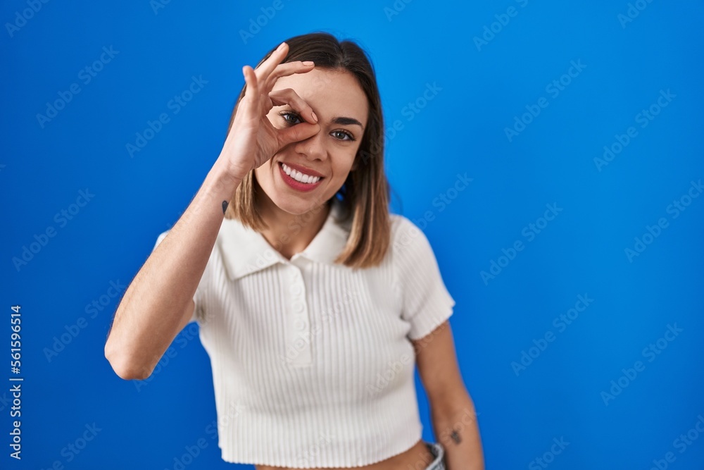 Hispanic woman standing over blue background doing ok gesture with hand smiling, eye looking through fingers with happy face.