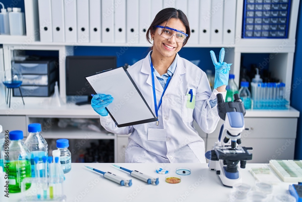 Hispanic young woman working at scientist laboratory smiling looking to the camera showing fingers doing victory sign. number two.