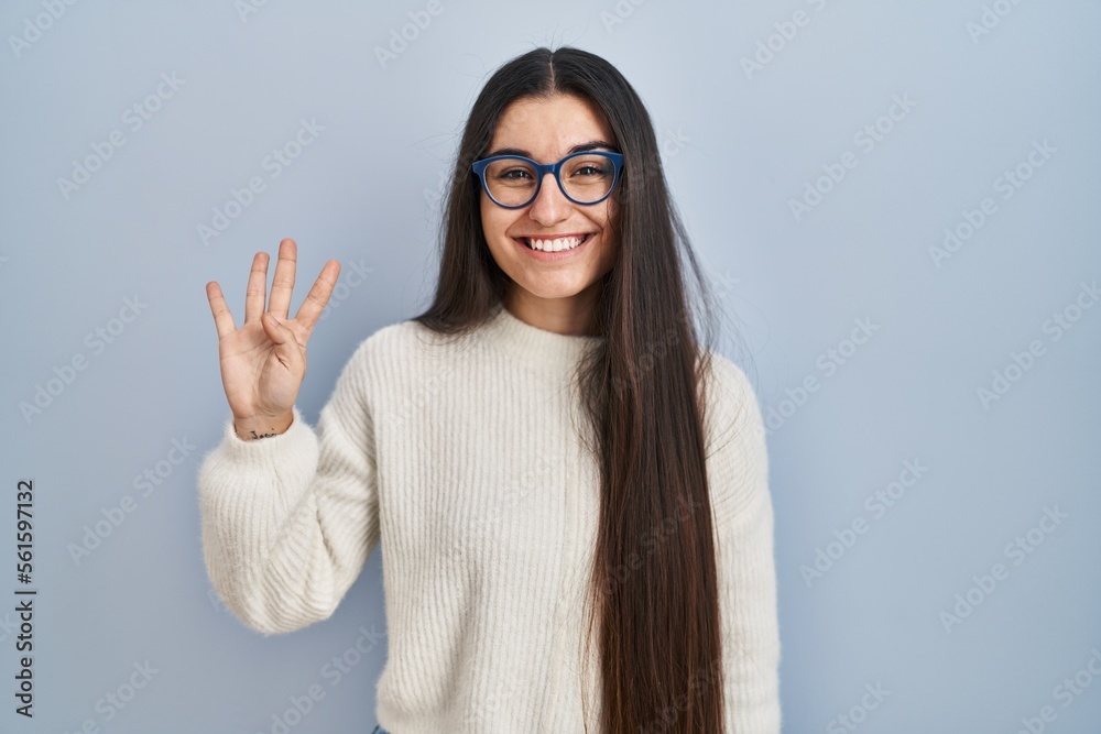 Young hispanic woman wearing casual sweater over blue background showing and pointing up with fingers number four while smiling confident and happy.
