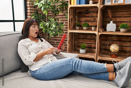 Young hispanic woman using touchpad sitting on the sofa scared and amazed with open mouth for surprise, disbelief face