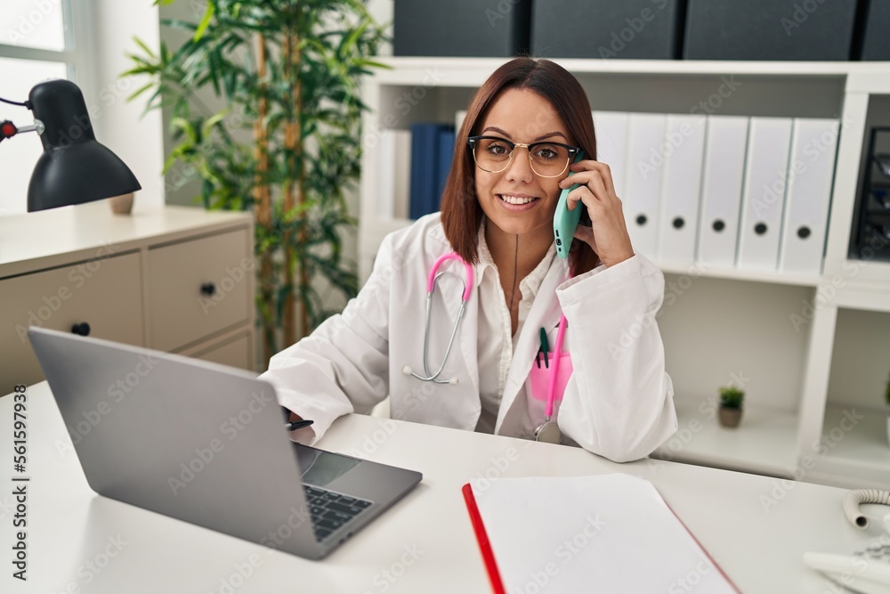 Young beautiful hispanic woman doctor talking on smartphone using laptop at clinic