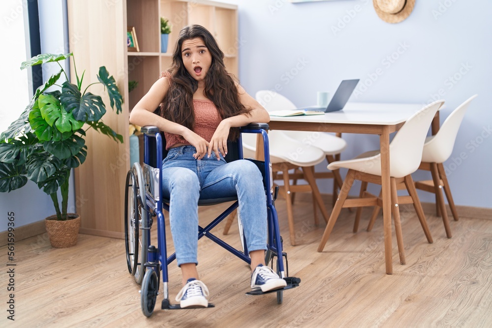 Young teenager girl sitting on wheelchair at the living room scared and amazed with open mouth for surprise, disbelief face Young teenager girl sitting on wheelchair at the living room scared and amazed with open mouth for surprise, disbelief face