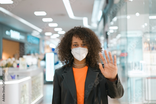 COVID-19 Pandemic Coronavirus. Portrait young African american girl wearing face mask ffp2 walk in the mall. Woman holding hand in stop sign, warning and preventing you from something bad