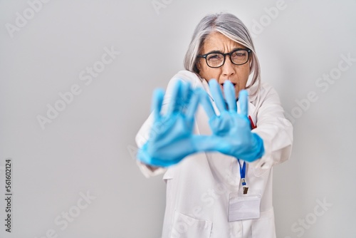 Middle age woman with grey hair wearing scientist robe afraid and terrified with fear expression stop gesture with hands  shouting in shock. panic concept.
