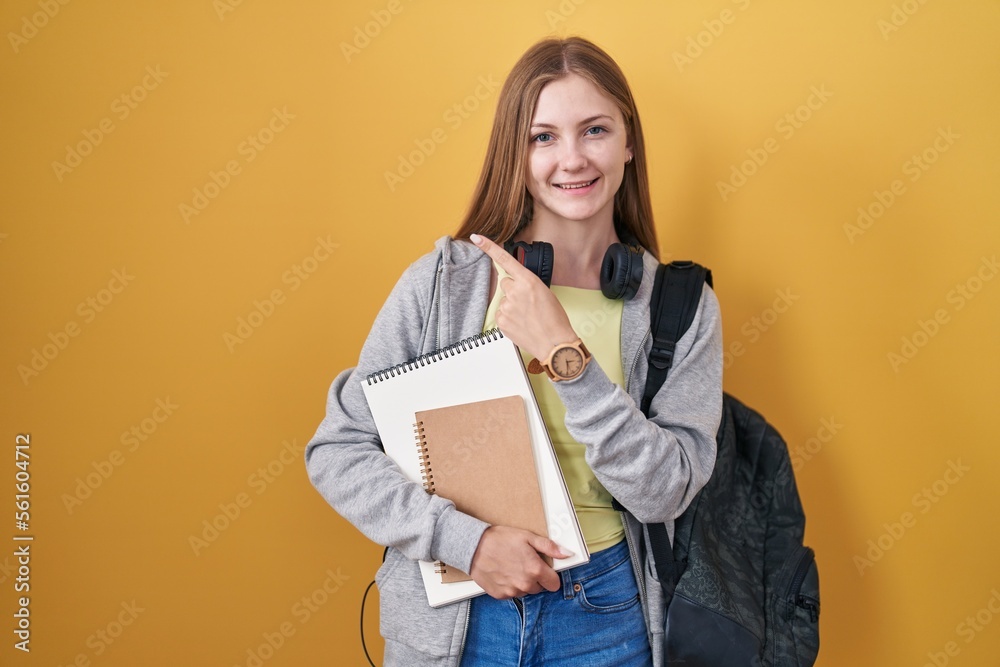 Fototapeta premium Young caucasian woman wearing student backpack and holding books with a big smile on face, pointing with hand finger to the side looking at the camera.