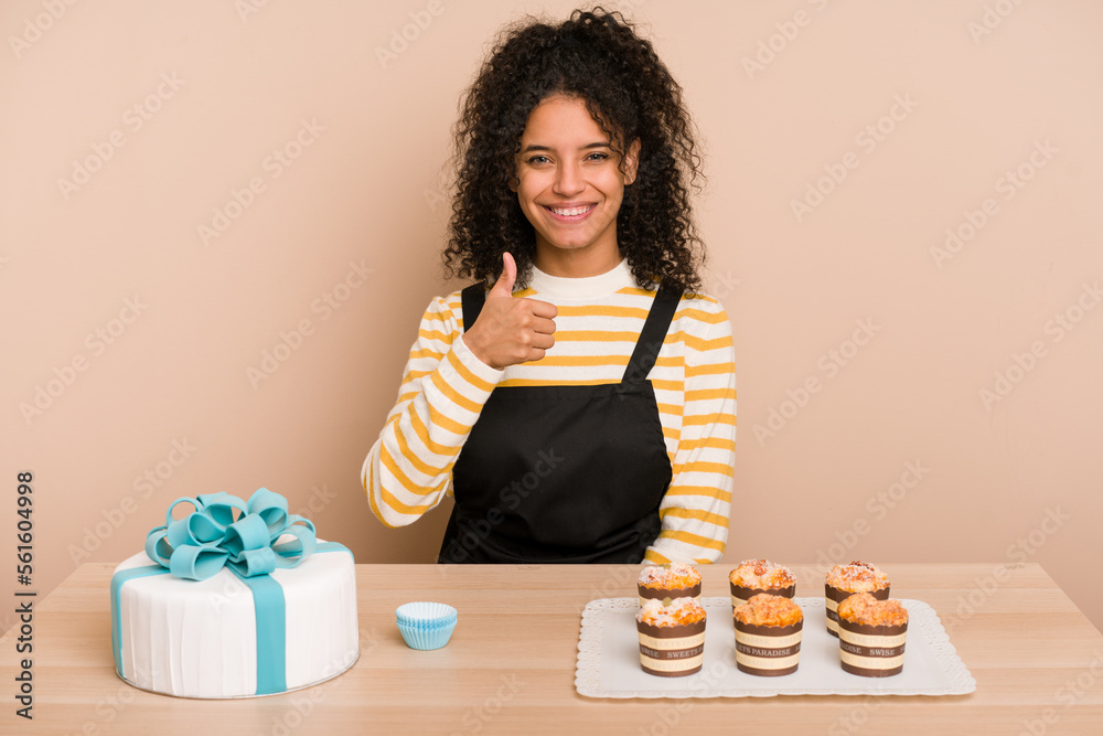 Young african american woman preparing a sweet cake and muffins on a table smiling and raising thumb up Young african american woman preparing a sweet cake and muffins on a table smiling and raising thumb up