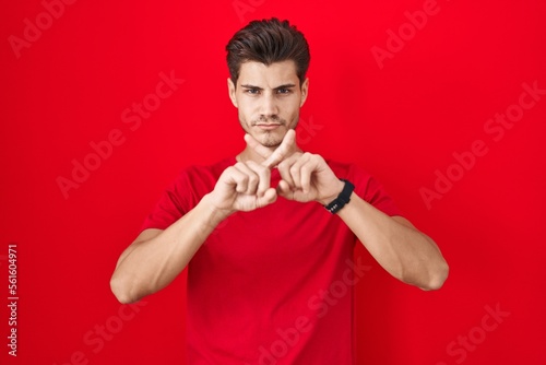 Young hispanic man standing over red background rejection expression crossing fingers doing negative sign
