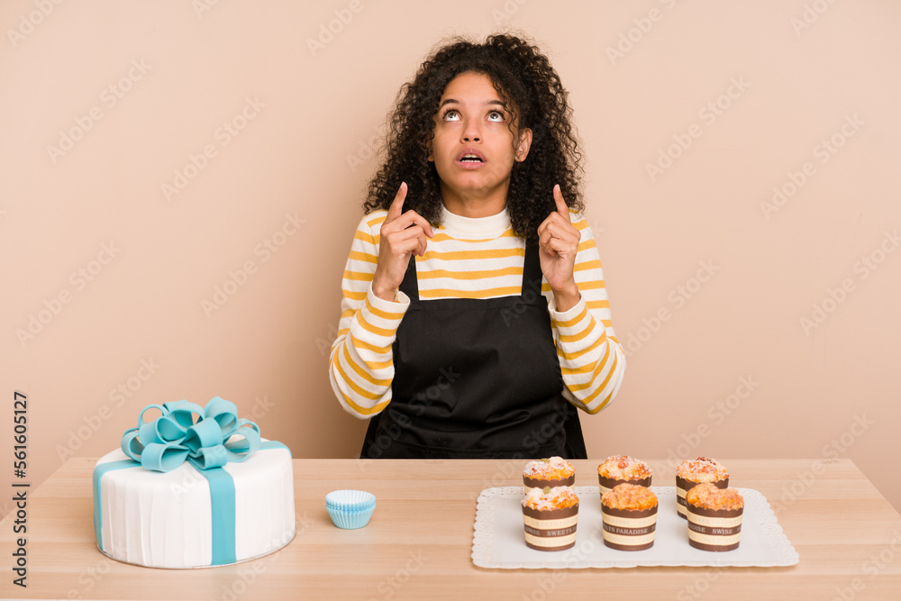 Young african american woman preparing a sweet cake and muffins on a table pointing upside with opened mouth. Young african american woman preparing a sweet cake and muffins on a table pointing upside with opened mouth.