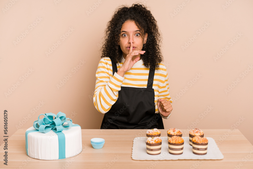Young african american woman preparing a sweet cake and muffins on a table keeping a secret or asking for silence.