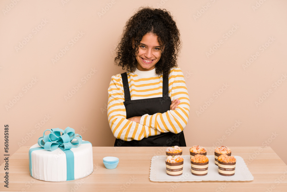 Young african american woman preparing a sweet cake and muffins on a table who feels confident, crossing arms with determination. Young african american woman preparing a sweet cake and muffins on a table who feels confident, crossing arms with determination.