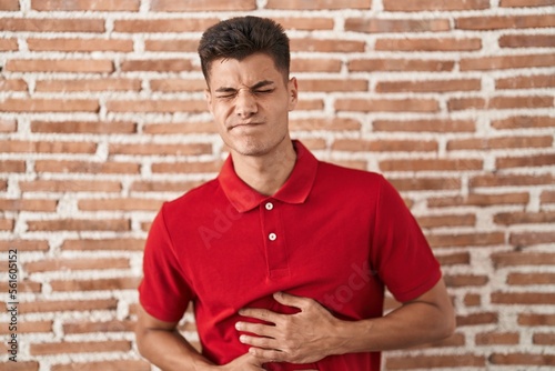 Young hispanic man standing over bricks wall with hand on stomach because nausea, painful disease feeling unwell. ache concept.