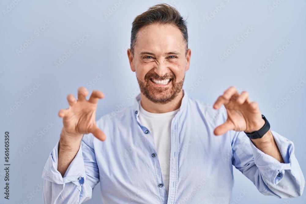 Middle age caucasian man standing over blue background smiling funny doing claw gesture as cat, aggressive and sexy expression Middle age caucasian man standing over blue background smiling funny doing claw gesture as cat, aggressive and sexy expression