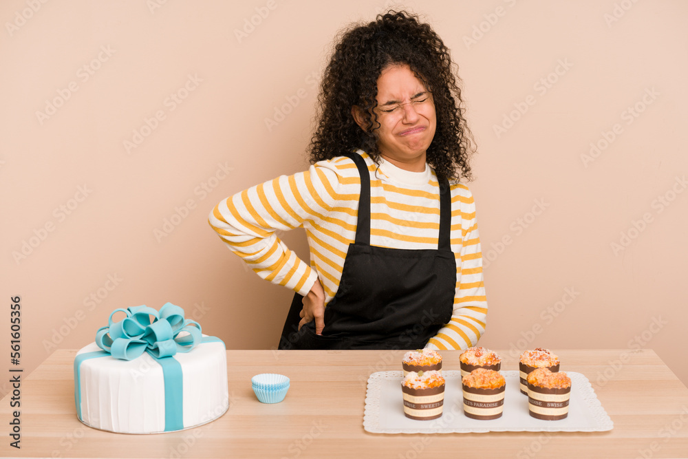 Young african american woman preparing a sweet cake and muffins on a table suffering a back pain. Young african american woman preparing a sweet cake and muffins on a table suffering a back pain.