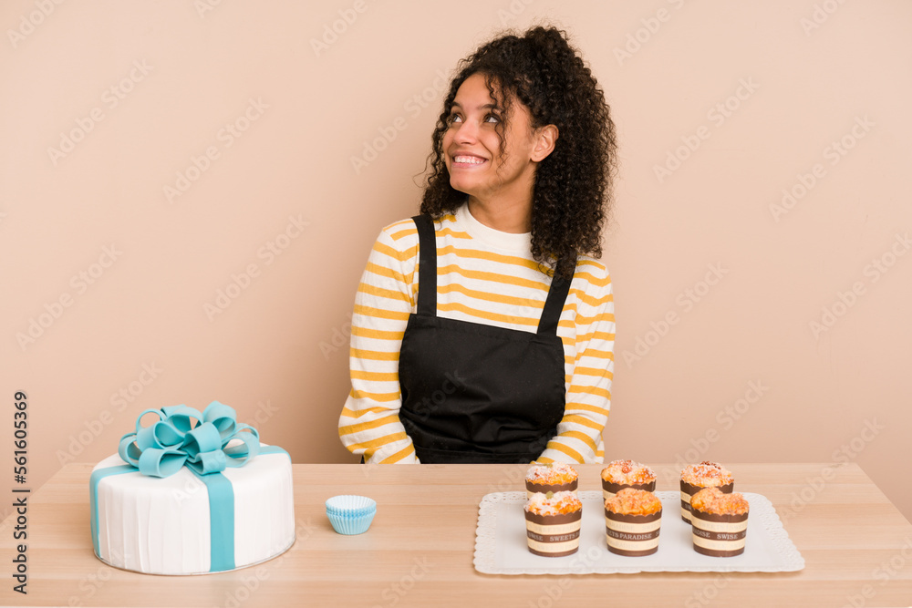 Young african american woman preparing a sweet cake and muffins on a table relaxed and happy laughing, neck stretched showing teeth. Young african american woman preparing a sweet cake and muffins on a table relaxed and happy laughing, neck stretched showing teeth.