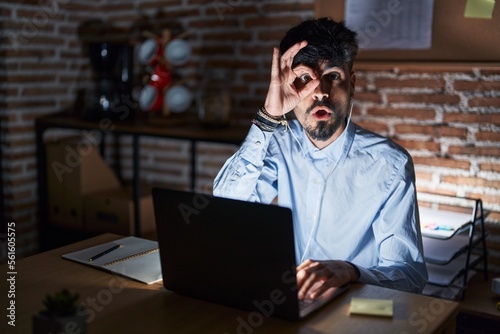 Young hispanic man with beard working at the office at night doing ok gesture shocked with surprised face, eye looking through fingers. unbelieving expression.