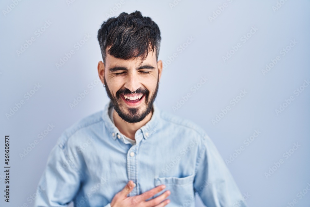 Obraz premium Young hispanic man with beard standing over blue background smiling and laughing hard out loud because funny crazy joke with hands on body.