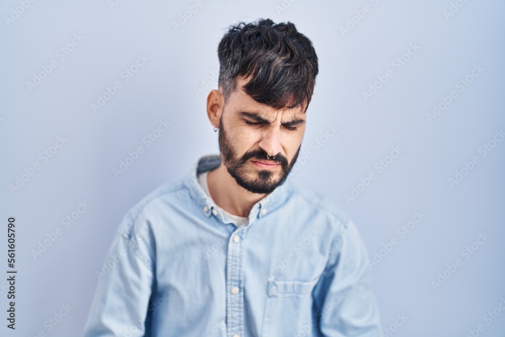 Young hispanic man with beard standing over blue background with hand on stomach because indigestion, painful illness feeling unwell. ache concept. Young hispanic man with beard standing over blue background with hand on stomach because indigestion, painful illness feeling unwell. ache concept.