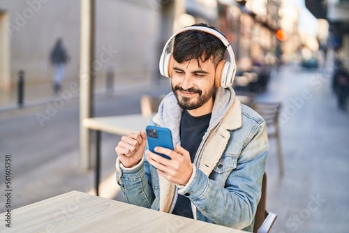 Young hispanic man listening to music sitting on table at coffee shop terrace