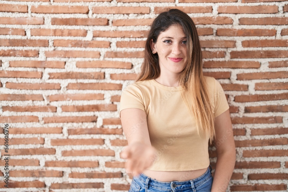 Young brunette woman standing over bricks wall smiling cheerful offering palm hand giving assistance and acceptance. Young brunette woman standing over bricks wall smiling cheerful offering palm hand giving assistance and acceptance.