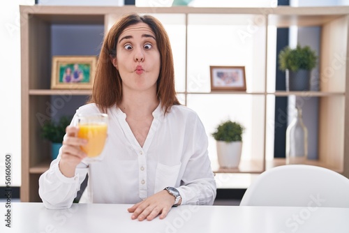 Brunette woman drinking glass of orange juice making fish face with lips, crazy and comical gesture. funny expression.