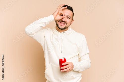 Young latin man holding a cola refreshment isolated excited keeping ok gesture on eye. © Asier
