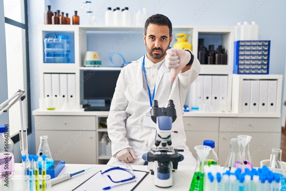 Young hispanic man with beard working at scientist laboratory looking unhappy and angry showing rejection and negative with thumbs down gesture. bad expression. Young hispanic man with beard working at scientist laboratory looking unhappy and angry showing rejection and negative with thumbs down gesture. bad expression.