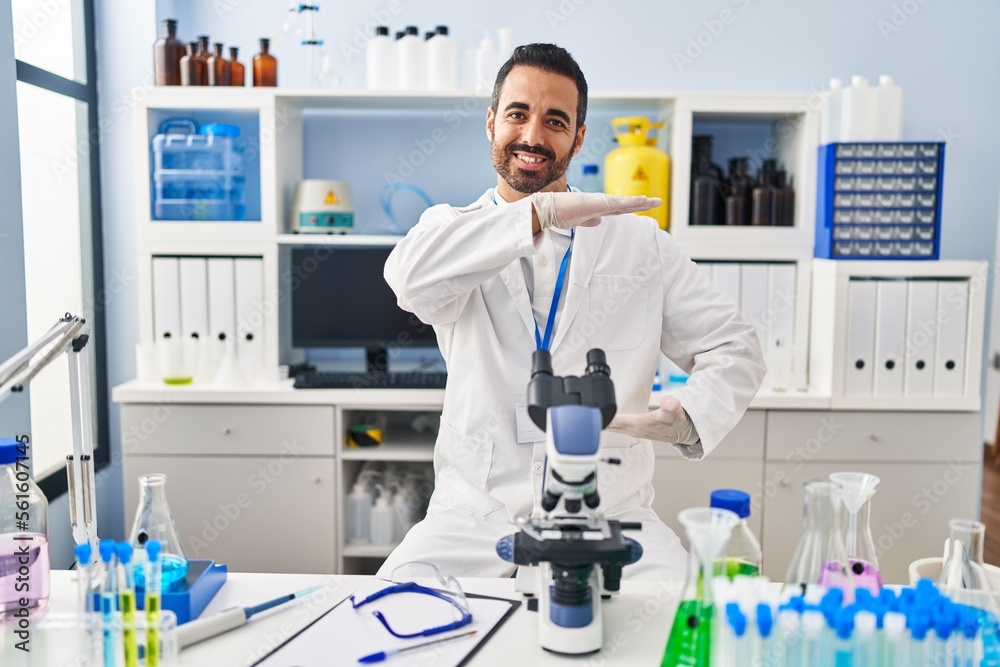 Young hispanic man with beard working at scientist laboratory gesturing with hands showing big and large size sign, measure symbol. smiling looking at the camera. measuring concept.