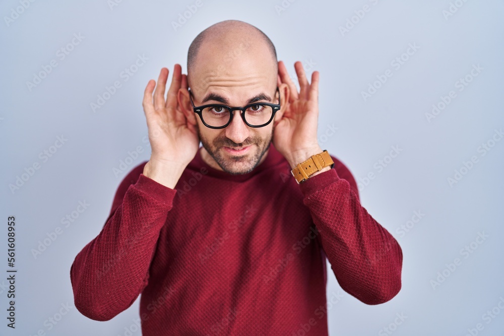 Young bald man with beard standing over white background wearing glasses trying to hear both hands on ear gesture, curious for gossip. hearing problem, deaf