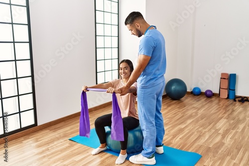 Latin man and woman wearing physiotherapist uniform having rehab session using fit ball and elastic band at rehab center