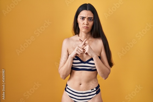 Young brunette woman wearing bikini over yellow background rejection expression crossing fingers doing negative sign