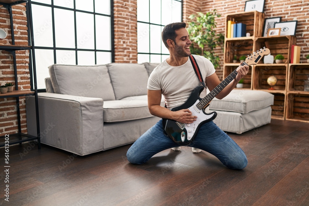Young hispanic man playing electrical guitar with knees on floor at home