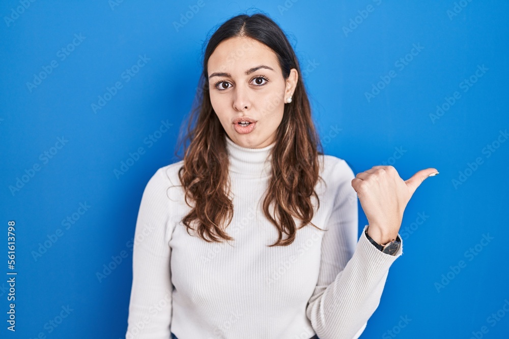 Young hispanic woman standing over blue background surprised pointing with hand finger to the side, open mouth amazed expression.