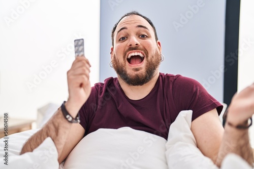 Plus size hispanic man with beard in the bed holding pills celebrating victory with happy smile and winner expression with raised hands