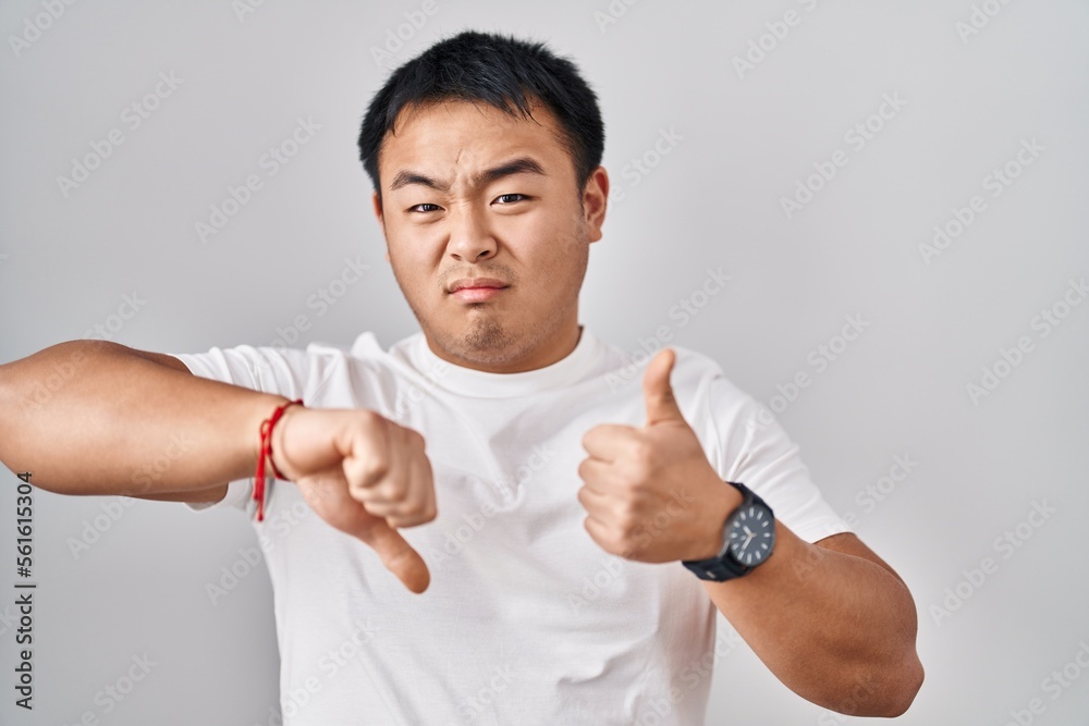 Young chinese man standing over white background doing thumbs up and down, disagreement and agreement expression. crazy conflict