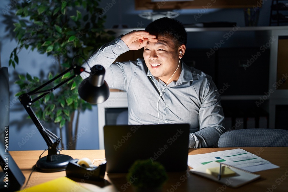 Young chinese man working using computer laptop at night very happy and smiling looking far away with hand over head. searching concept. Young chinese man working using computer laptop at night very happy and smiling looking far away with hand over head. searching concept.
