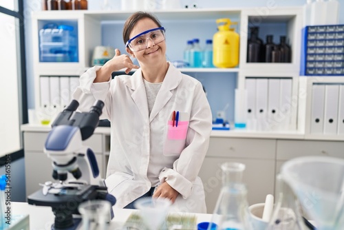 Hispanic girl with down syndrome working at scientist laboratory smiling doing phone gesture with hand and fingers like talking on the telephone. communicating concepts.