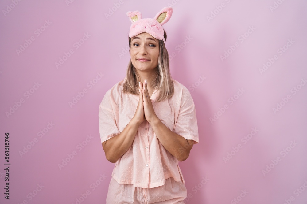 Blonde caucasian woman wearing sleep mask and pajama praying with hands together asking for forgiveness smiling confident. Blonde caucasian woman wearing sleep mask and pajama praying with hands together asking for forgiveness smiling confident.