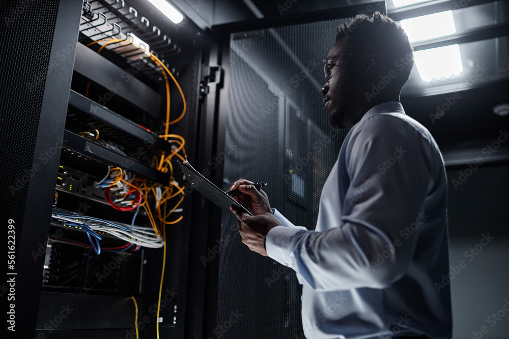 Backlit side view portrait of black man working with server cabinet in data center and taking notes on clipboard, copy space - obrazy, fototapety, plakaty 