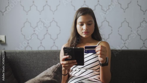 Happy young brown haired girl sitting on sofa couch at home holding smartphone and credit card in hand doing online shopping payment, phone banking, transaction. E-banking app service, Finance economy photo