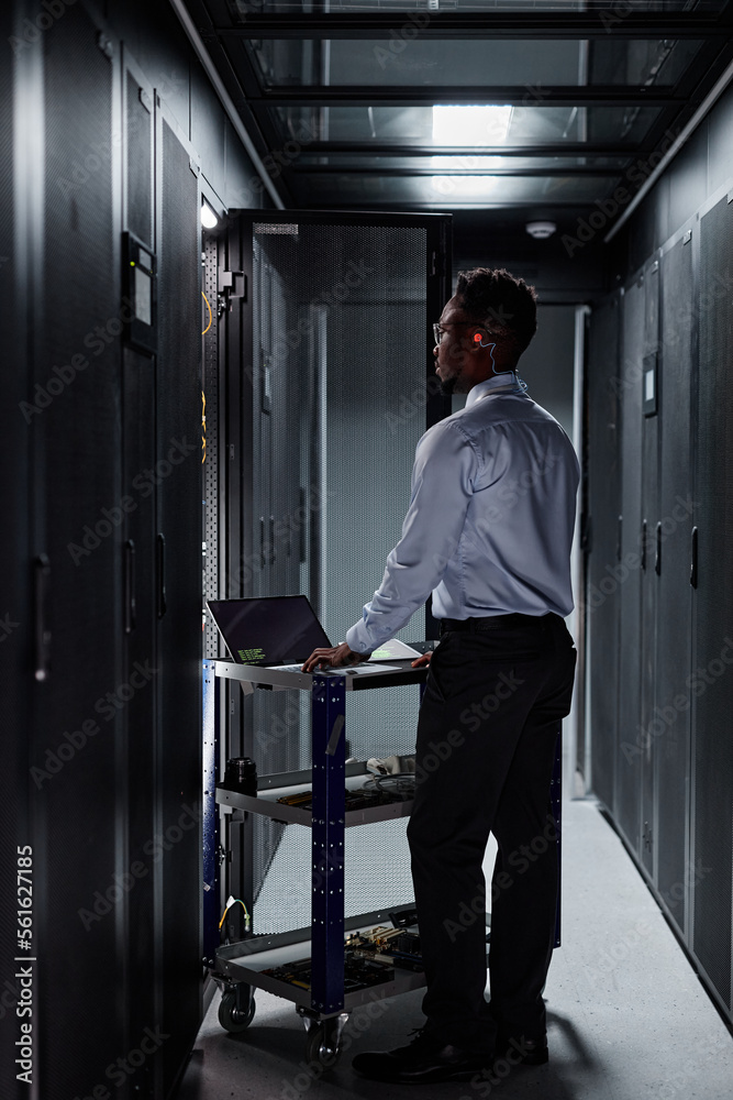 Vertical full length portrait of black man as network engineer using laptop and setting up servers in data center Vertical full length portrait of black man as network engineer using laptop and setting up servers in data center
