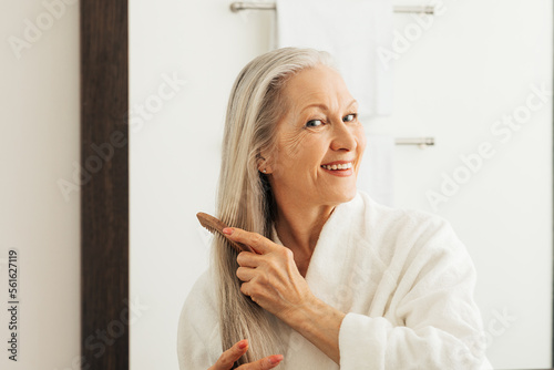 Canvas Print Cheerful woman combing her long hair with a wood comb in front of a bathroom mir