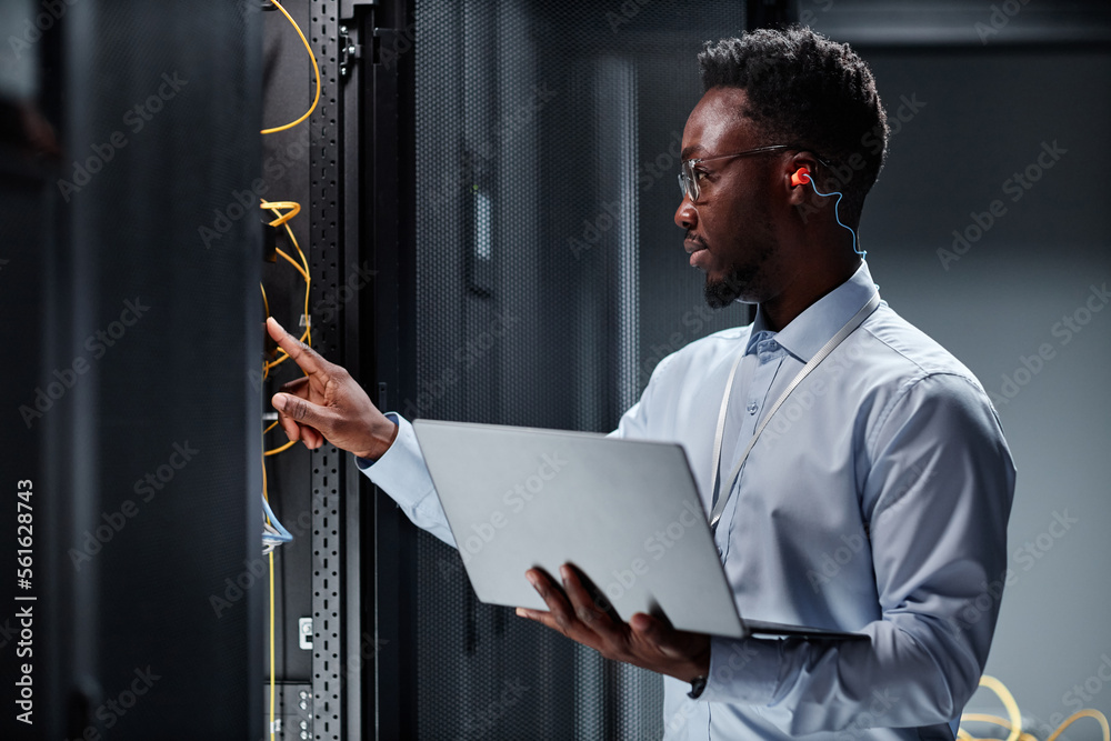 © Seventyfour - Side view portrait of young black man as network engineer working with servers in data center and holding laptop © Seventyfour - Side view portrait of young black man as network engineer working with servers in data center and holding laptop