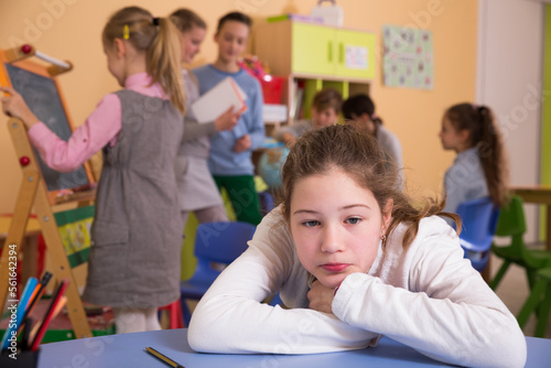 Upset girl sitting at table in schoolroom during break on background with other pupils