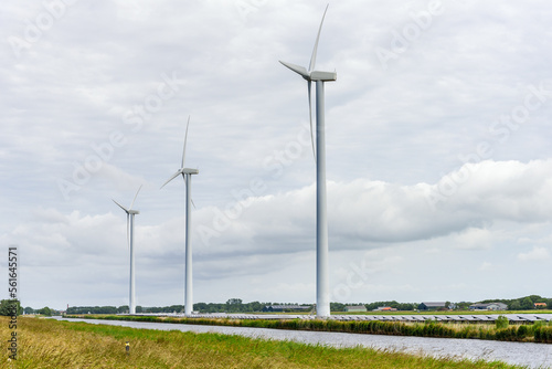 Wind turbines along a canal in the countryside of Netherlands on a cloudy summer day. Rows of solar panels are at the foot of the turbines.