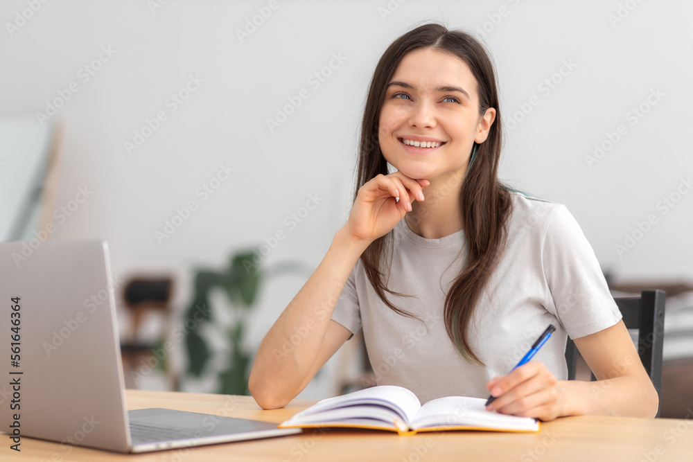 Young woman student looks dreamily away, thinking about of something good sitting at a table at home. Distance learning, online education concept Young woman student looks dreamily away, thinking about of something good sitting at a table at home. Distance learning, online education concept