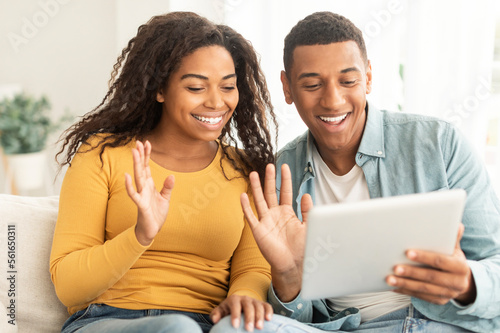 Cheerful young african american guy and lady waving hands at tablet webcam, greeting, say hello on sofa