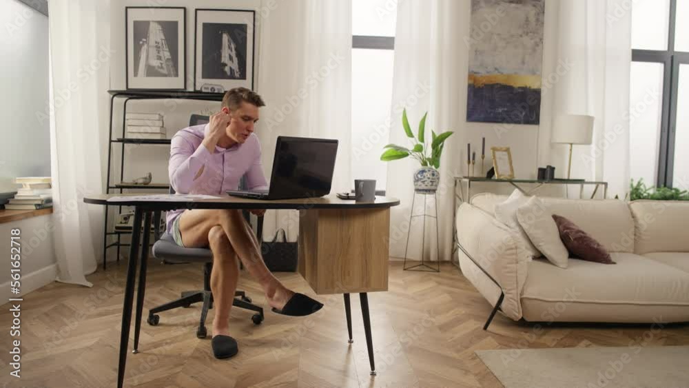 Businessman on zoom meeting from home office. A man in shorts and a shirt works remotely from home. It is convenient to work online at the computer in slippers. High quality 4k footage Businessman on zoom meeting from home office. A man in shorts and a shirt works remotely from home. It is convenient to work online at the computer in slippers. High quality 4k footage