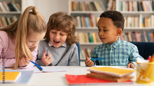 School time. Multiethnic kids writing in notebooks and smiling, talking with each other and laughing, sitting at table