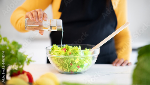 Young african american woman in apron preparing salad, pours oil at table with fresh vegetables