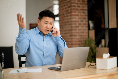 Displeased mature asian man talking on cellphone while working on laptop, having problem with business communication